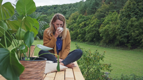 A young woman in sweater drinking coffee and sitting on floor outdoors on patio with forest at background.