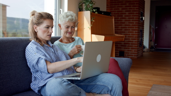 A senior mother having coffee with adult daughter indoors at home, sitting, talking and using laptop.