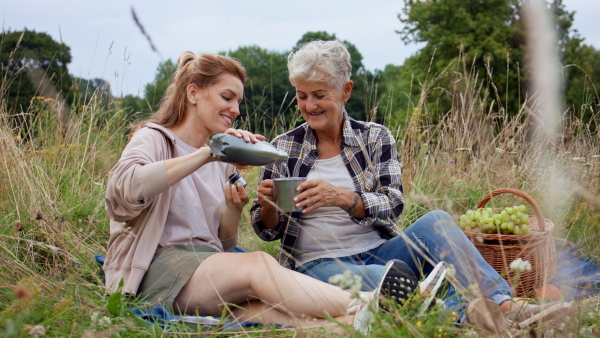 A happy senior mother and adult daughter sitting and having picnic outdoors in nature.