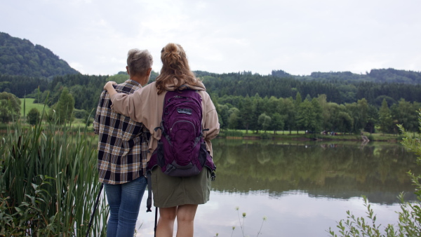 A rear view of senior mother embracing with adult daughter when standing by lake outdoors in nature