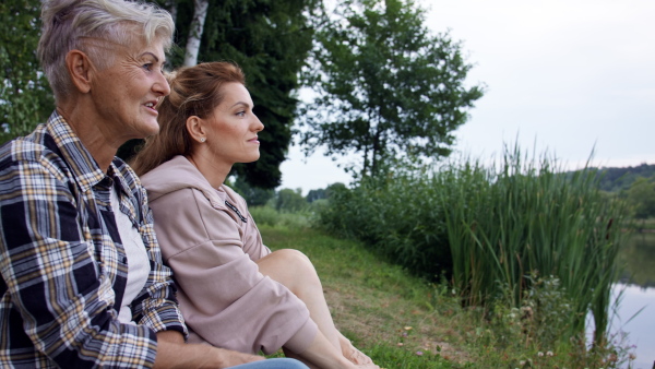 A profile view of happy senior mother hiker sitting with adult daughter by lake outdoors in nature