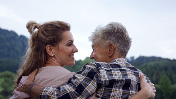 A rear view of senior mother embracing with adult daughter when standing by lake outdoors in nature