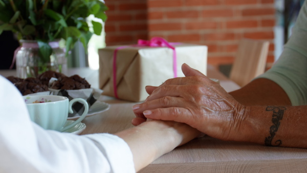 A close up of senior mother holding hand of adult daughter when having coffee together indoors at home