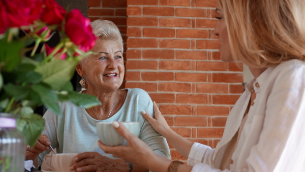 A happy senior mother talking with adult daughter when sitting and having coffee indoors at home.