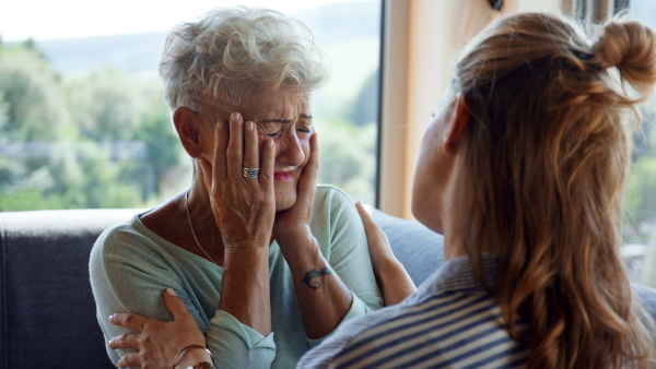 An adult daughter consoling unhappy senior mother and supporting her in struggle indoors at home.