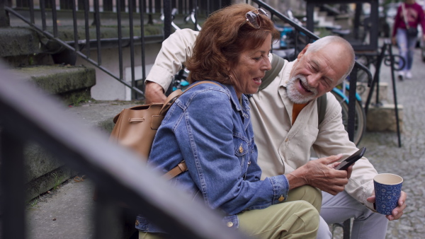 A happy senior couple tourists sitting on stairs and having take away coffee outdoors in town