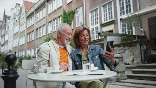 A happy senior couple tourists sitting and using smartphone outdoors in town.