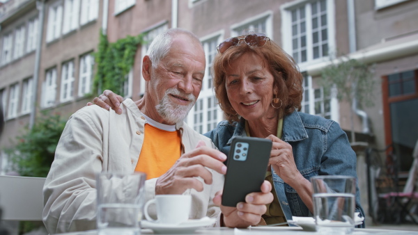 A happy senior couple tourists sitting and using smartphone outdoors in town.