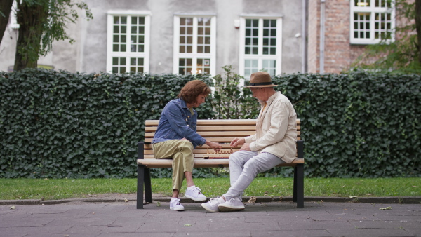 A happy senior couple sitting on bench and playing chess outdoors in park.