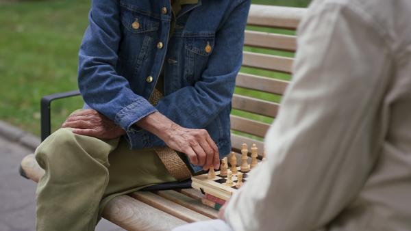 A happy senior couple sitting on bench and playing chess outdoors in park.