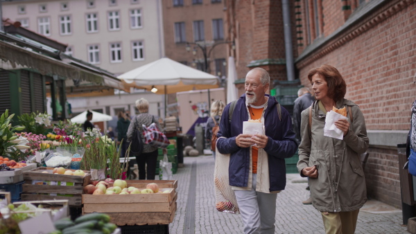 A happy senior couple tourists with snack in town on outdoor market.