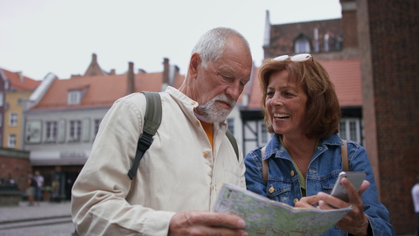 A happy senior couple tourists using smartphone and map outdoors in historic town