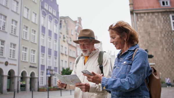 A happy senior couple tourists using smartphone and map outdoors in historic town