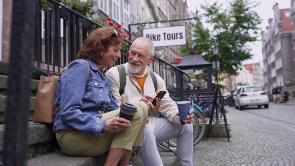 A happy senior couple tourists sitting on stairs and having take away coffee outdoors in town