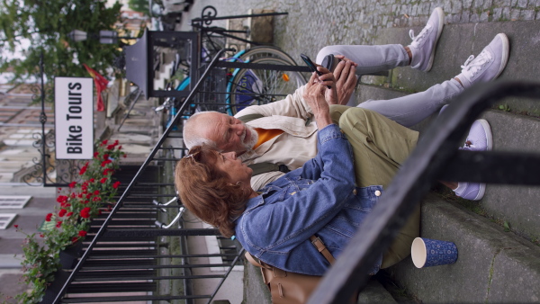 A happy senior couple tourists sitting on stairs and having take away coffee outdoors in town