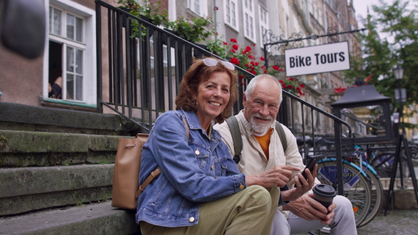 A happy senior couple tourists sitting on stairs and having take away coffee outdoors in town