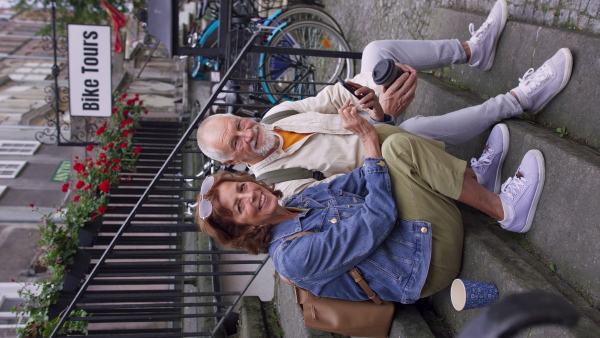 A happy senior couple tourists sitting on stairs and having take away coffee outdoors in town