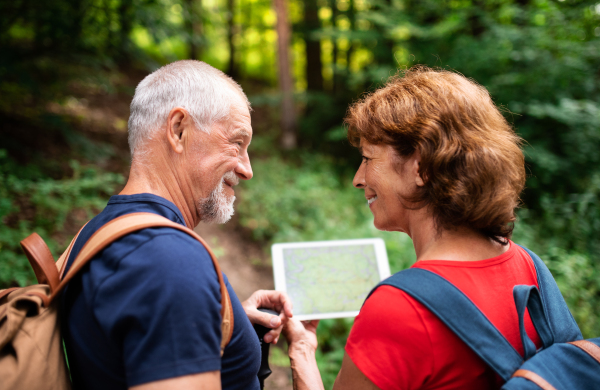 A senior tourist couple with backpacks on a walk in forest in nature, using map on tablet.