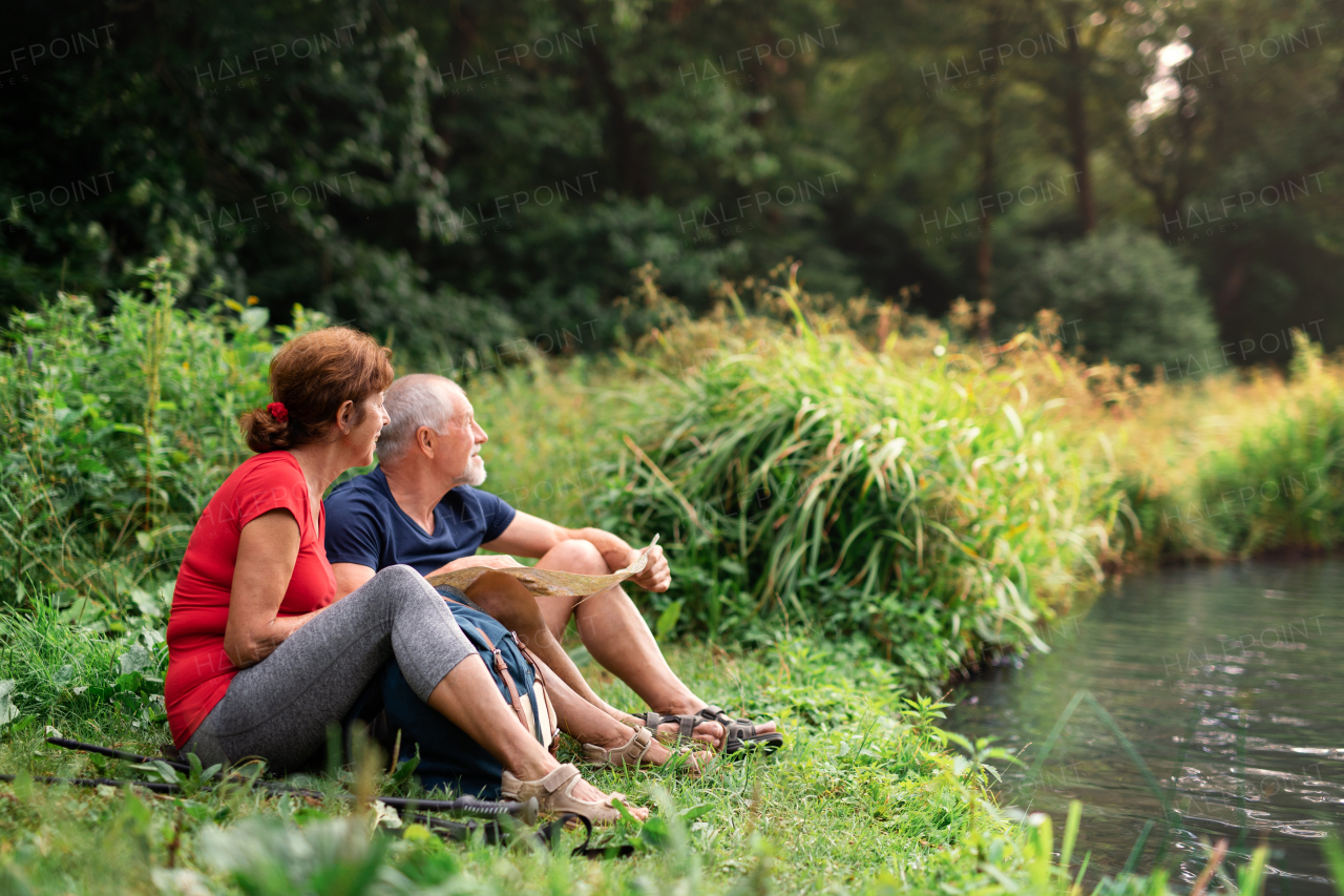 Senior tourist couple with map on a walk in nature, sitting by lake and resting.