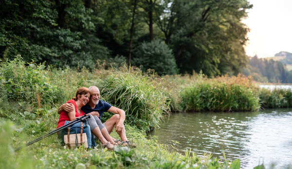 Senior tourist couple with backpacks on a walk in nature, sitting by lake and resting.