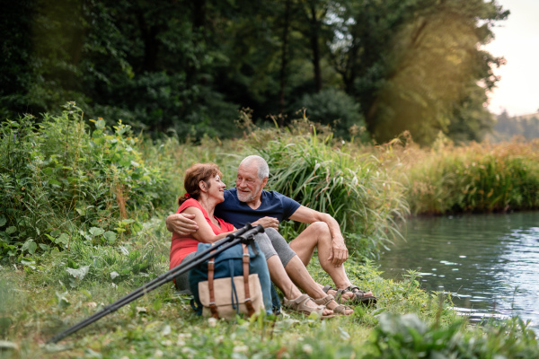 Senior tourist couple with backpacks on a walk in nature, sitting by lake and resting.