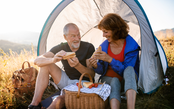 A senior tourist couple with shelter sitting in nature at sunset, eating sandwiches.