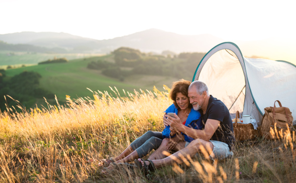 A senior tourist couple with smartphone sitting in nature at sunset, taking selfie.