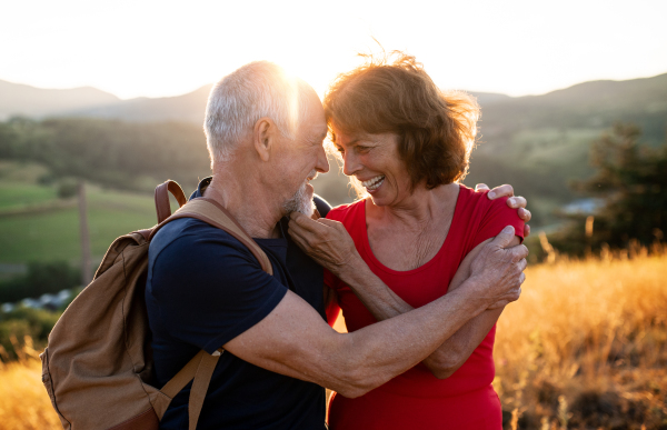 A senior tourist couple travellers with backpacks hiking in nature at sunset, hugging when resting.