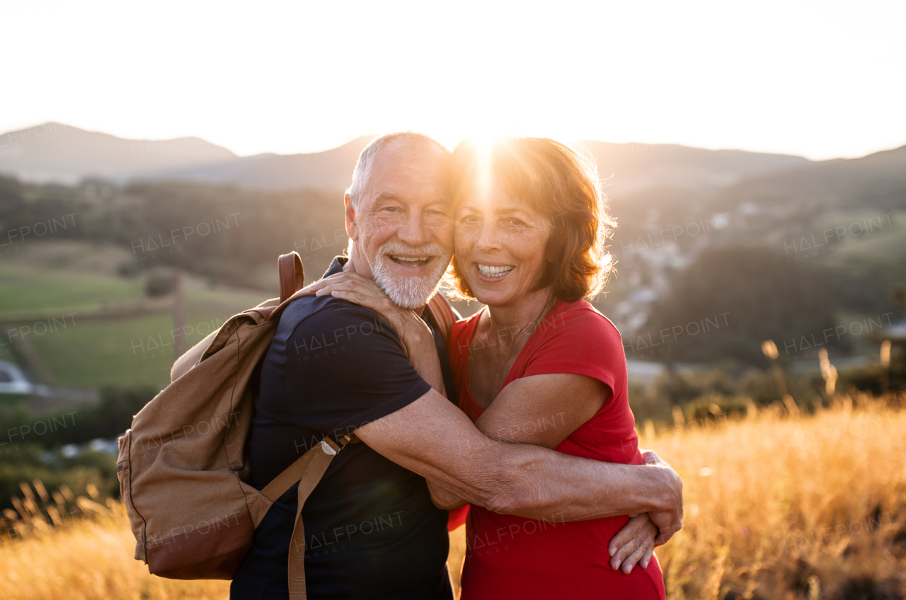 A senior tourist couple travellers with backpacks hiking in nature at sunset, hugging when resting.