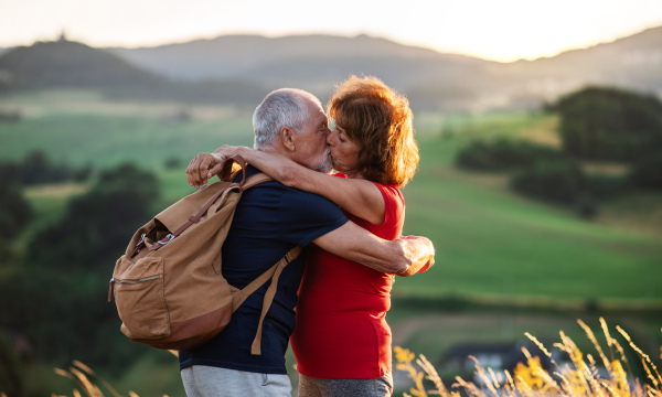 A senior tourist couple travellers with backpacks hiking in nature at sunset, kissing when resting. Copy space.