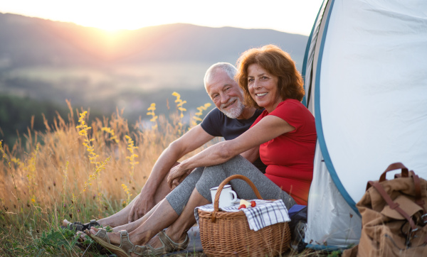 A senior tourist couple with picnic basket sitting in nature at sunset, resting.