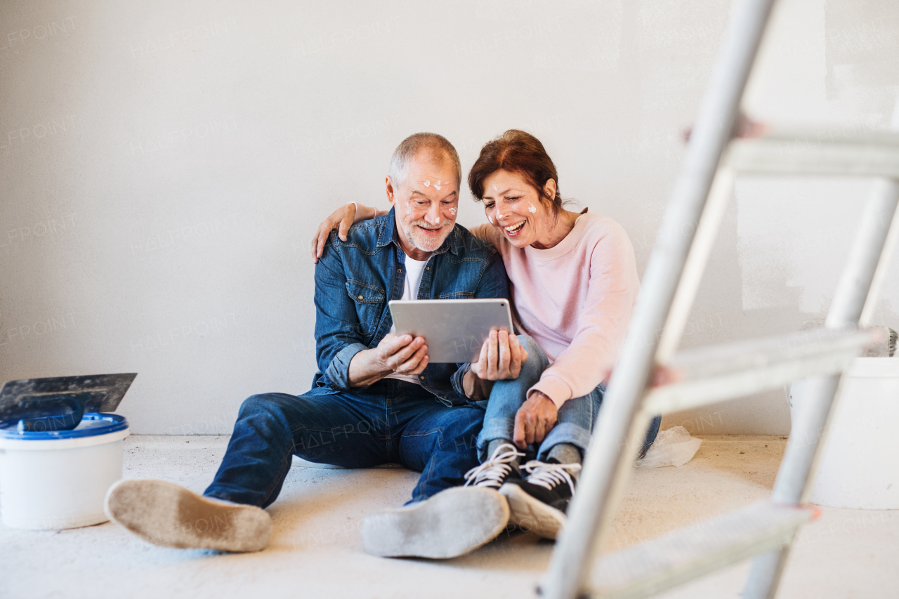 Happy senior couple painting walls in new home, using tablet. Relocation concept.