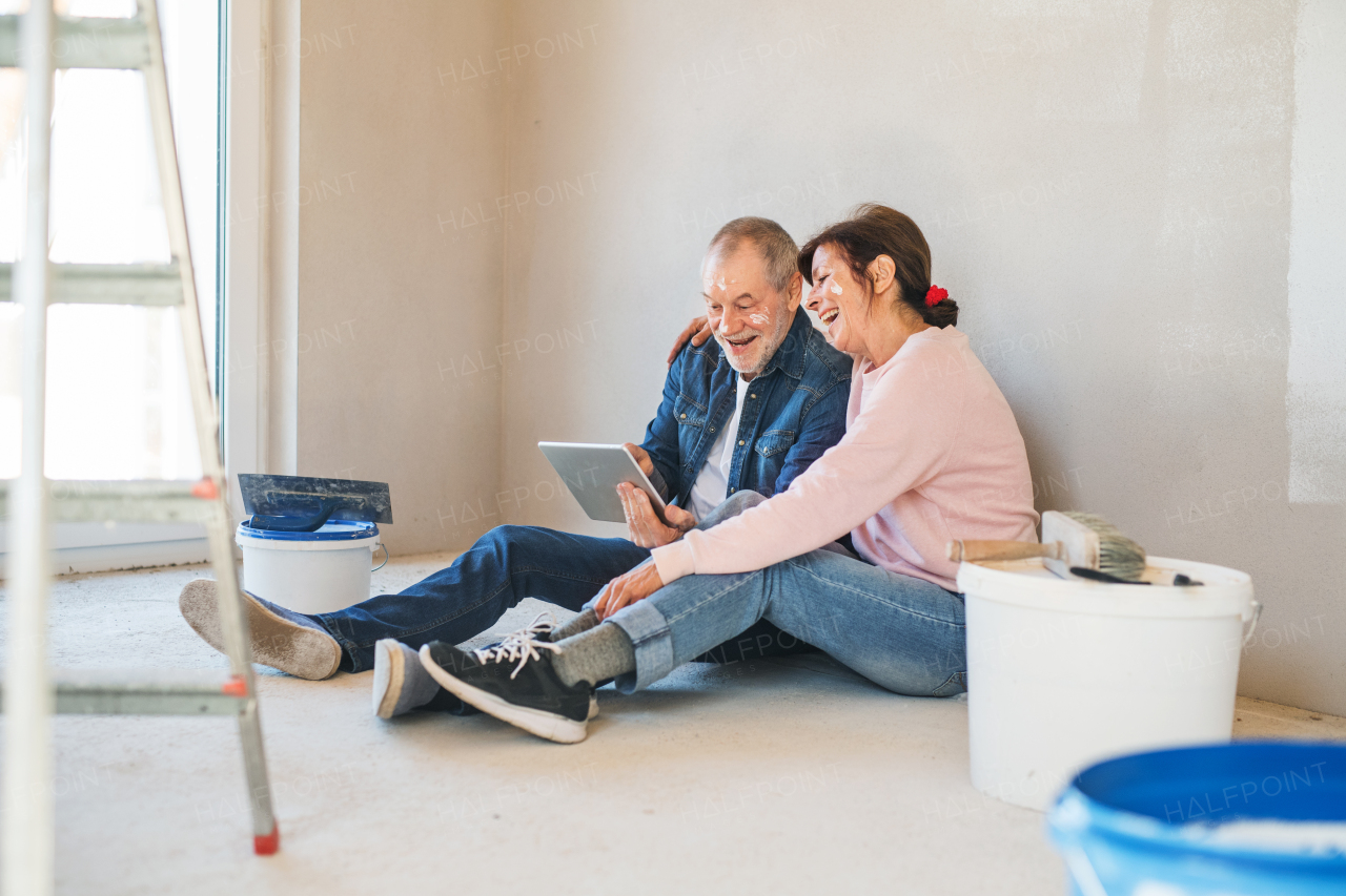 Happy senior couple painting walls in new home, using tablet. Relocation concept.