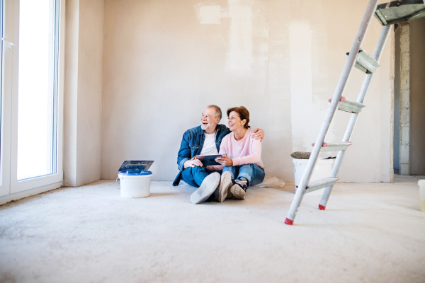 Happy senior couple painting walls in new home, using tablet. Relocation concept.