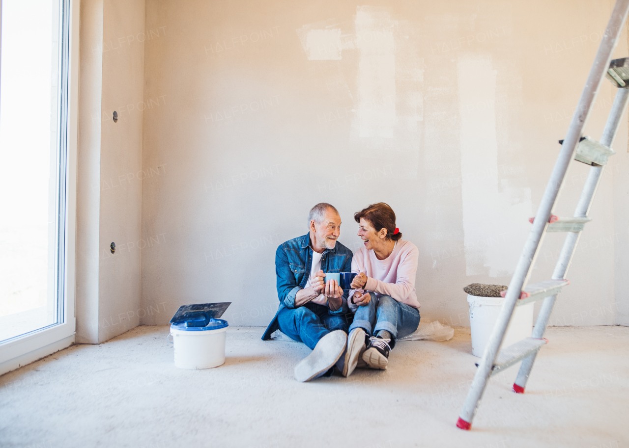 Portrait of cheerful senior couple with coffee or tea painting walls in new home, resting. Relocation concept.