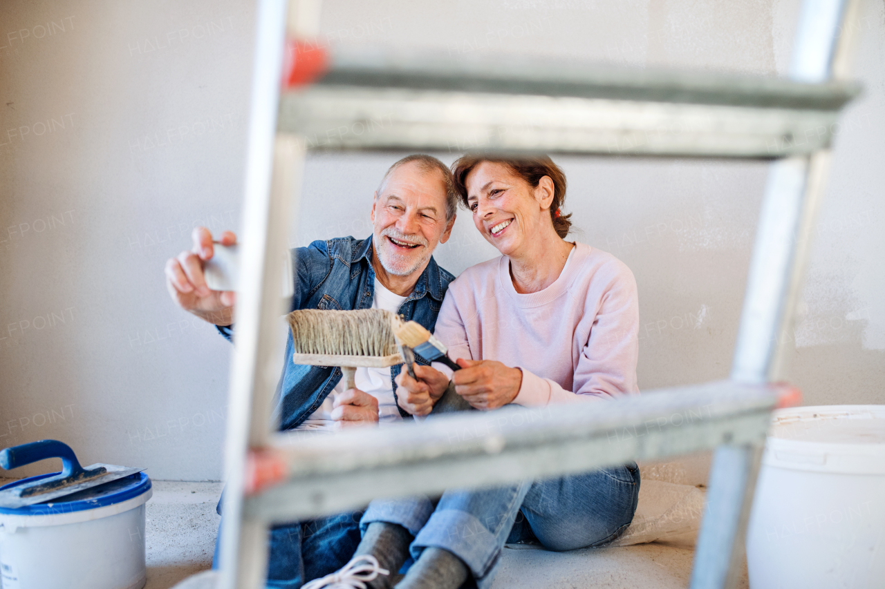 Senior couple taking selfie with smartphone when painting walls in new home, relocation concept.