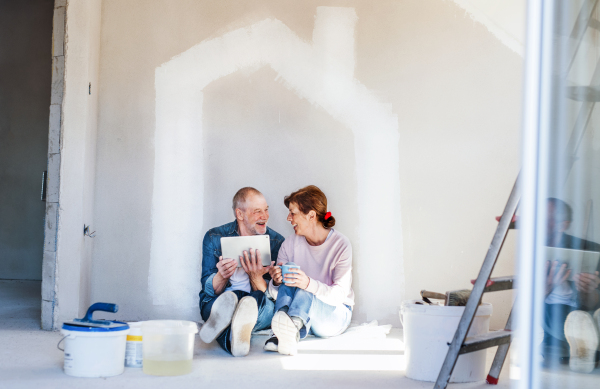 Happy senior couple painting walls in new home, using tablet. Relocation concept.