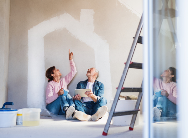 Happy senior couple painting walls in new home, using tablet. Relocation concept.