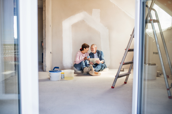 Happy senior couple painting walls in new home, using tablet. Relocation concept.