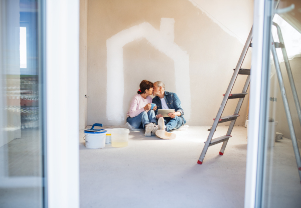 Happy senior couple with tablet painting walls in new home, kissing. Relocation concept.