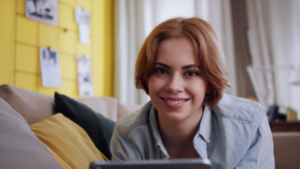 A cheerful teenager laying on sofa, using tablet and looking at camera. Social networks concept.