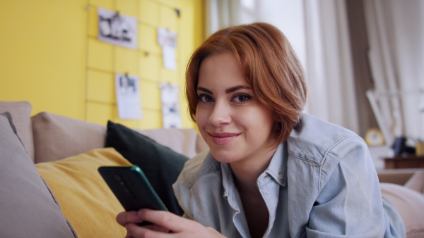 A cheerful teenager laying on sofa, using smartphone and looking at camera. Social networks concept.