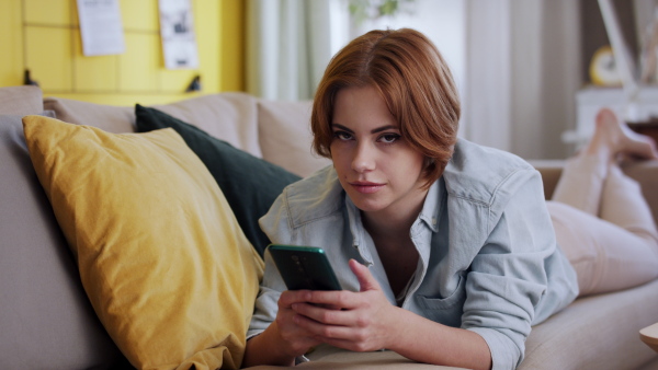 A cheerful teenager laying on sofa, using smartphone and looking at camera. Social networks concept.