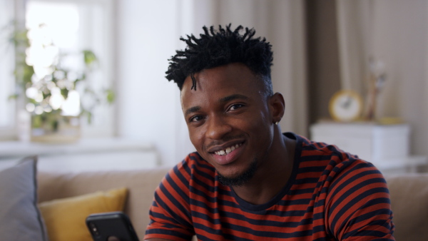 A cheerful afro-american teenager sitting on sofa, using smartphone and looking at camera. Social networks concept.