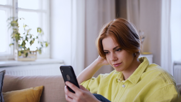 A cheerful teenager sitting on sofa, using smartphone and looking at camera. Social networks concept.