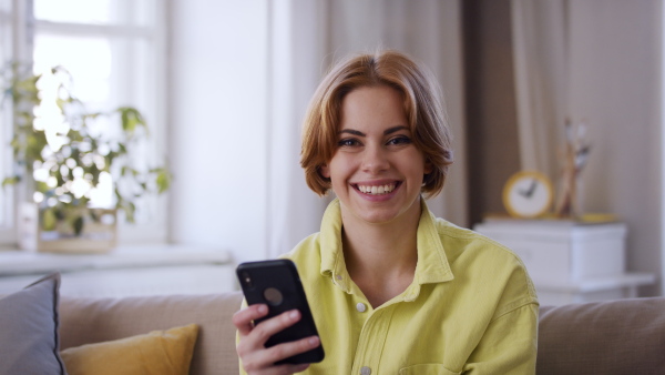 A cheerful teenager sitting on sofa, using smartphone and looking at camera. Social networks concept.