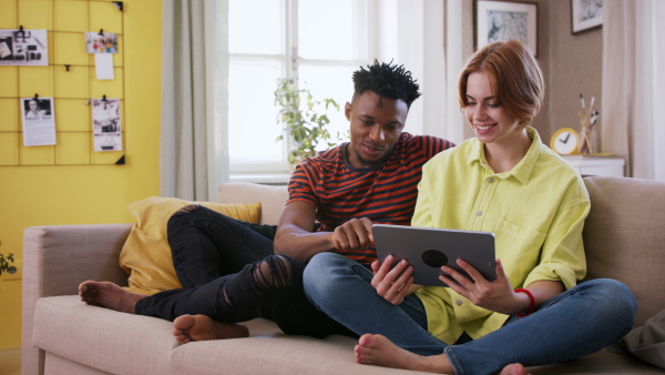 A cheerful teenagers sitting on sofa, using tablet, talking and looking at camera. Social networks concept.