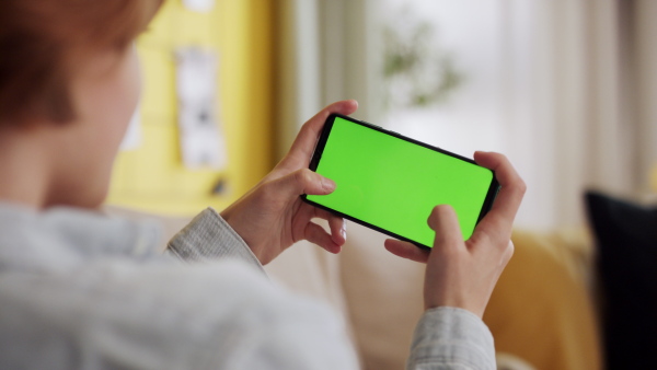 A rear view of teenager sitting on sofa and playing games smartphone with green screen.