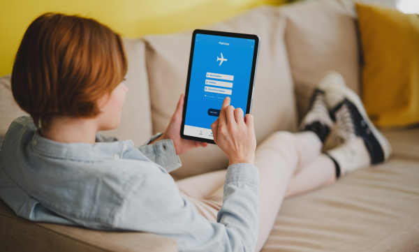 A rear view of young woman using travel app on tablet, lying on sofa at home,