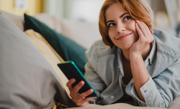 Portrait of happy young woman using smartphone, lying on sofa at home, looking aside, social networks concept.